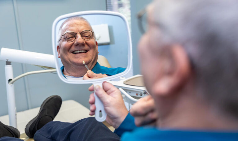 Senior dental patient smiling while examining a dental crown match in a handheld mirror